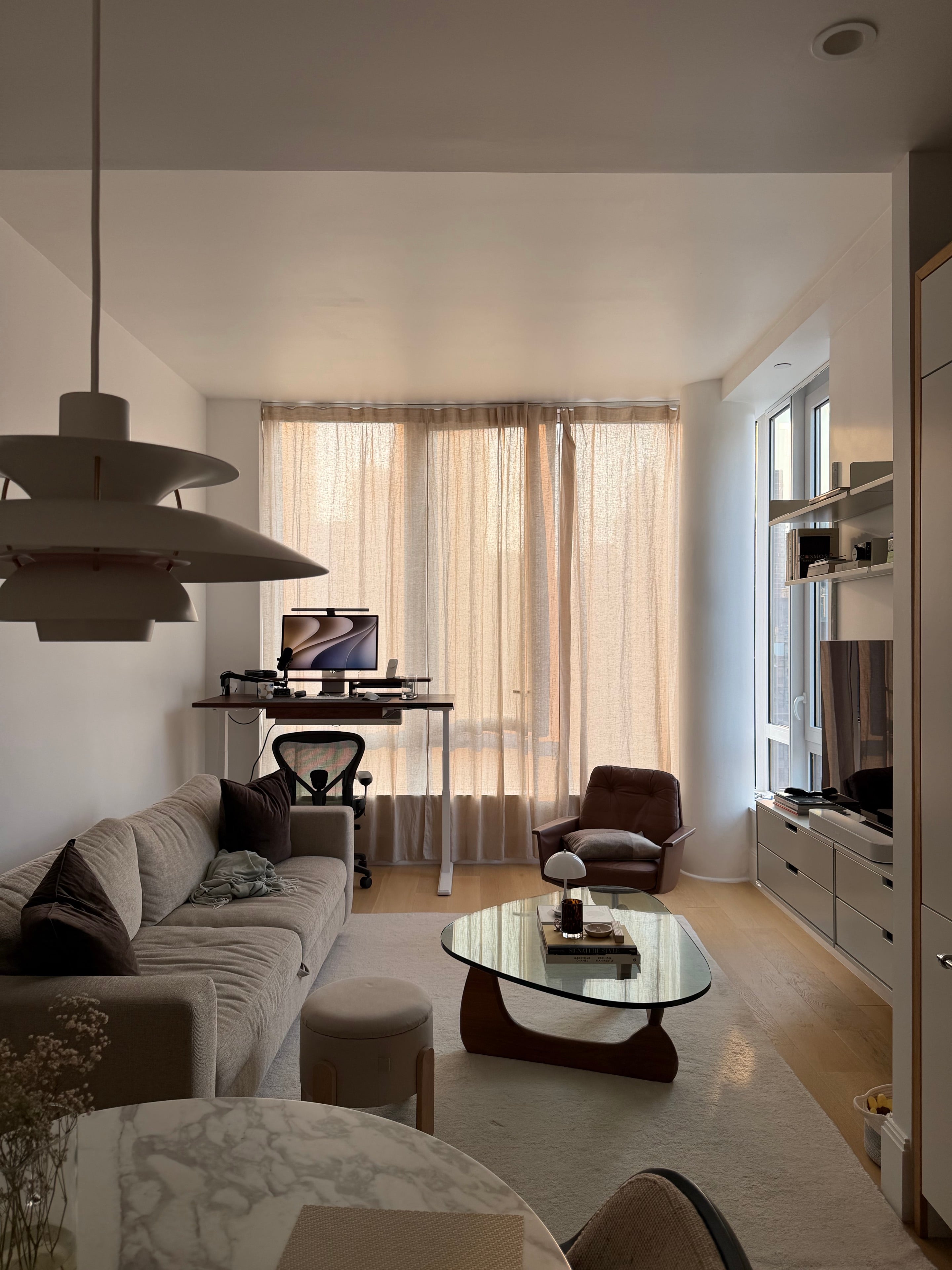 The desk in the living room, with a sofa, lounge chair, glass coffee table, and linen curtains